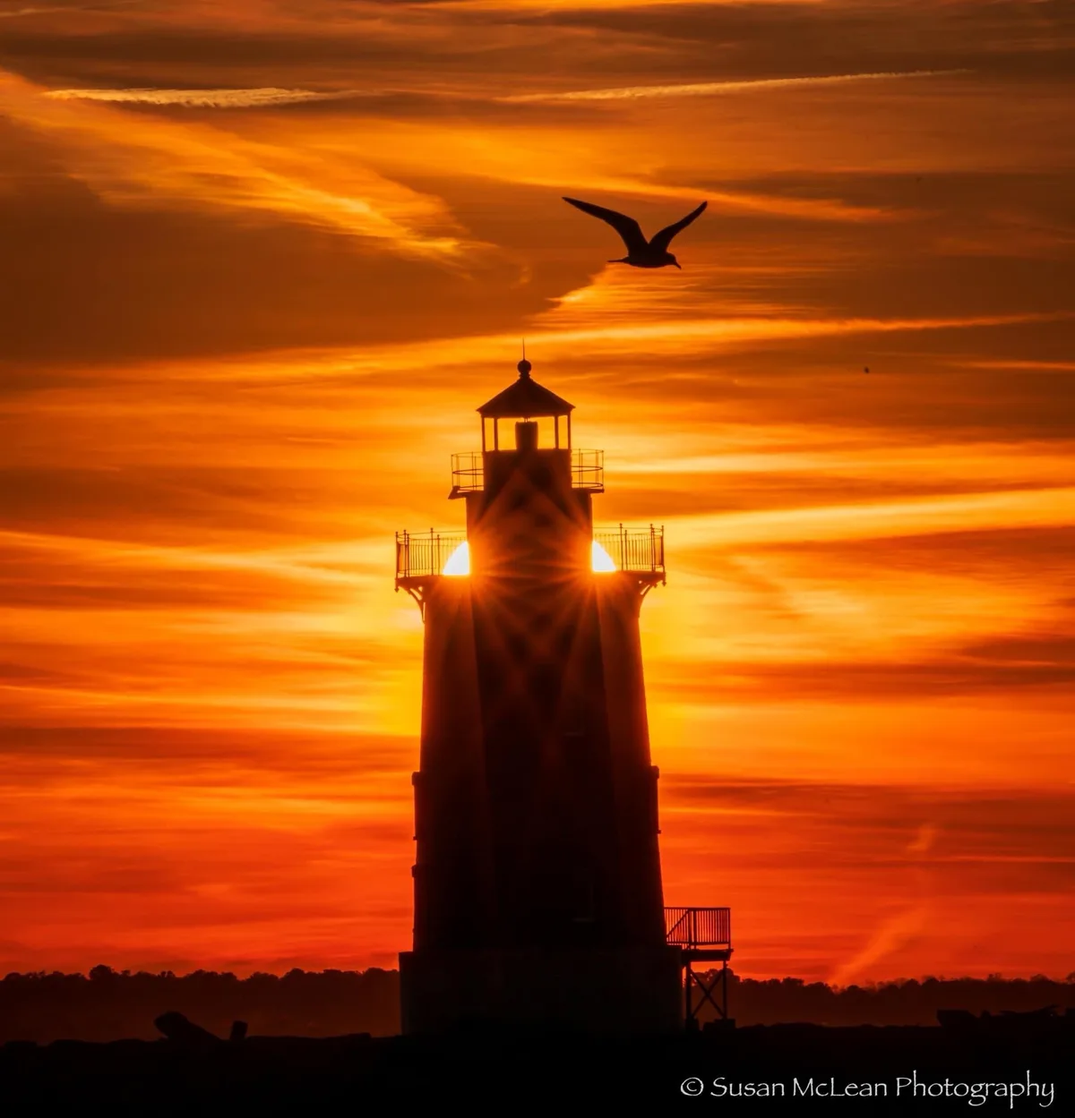 A lighthouse stands silhouetted against a vibrant orange sunset sky, with the sun's rays bursting from its lantern room. A…
