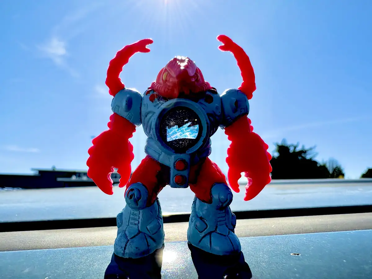 A red and blue toy robot stands outdoors under a bright sun, its arms raised in a triumphant pose. The robot's chest…