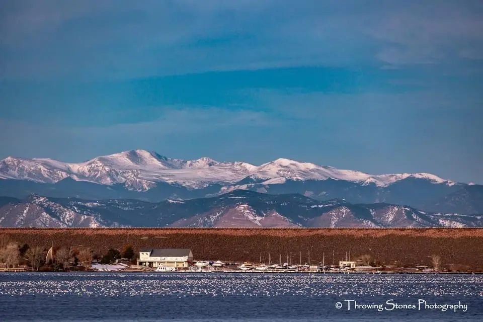 A scenic landscape photo shows a body of water in the foreground with a small dock and buildings, snow-capped mountains in…