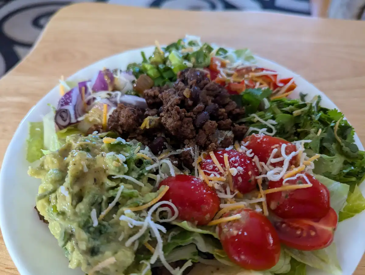 A close-up photo of a white plate holding a taco salad with lettuce, guacamole, cherry tomatoes, shredded cheese, black…