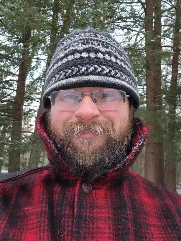 A man with glasses and a patterned knit hat stands in a snowy forest wearing a red and black plaid coat. His beard is…