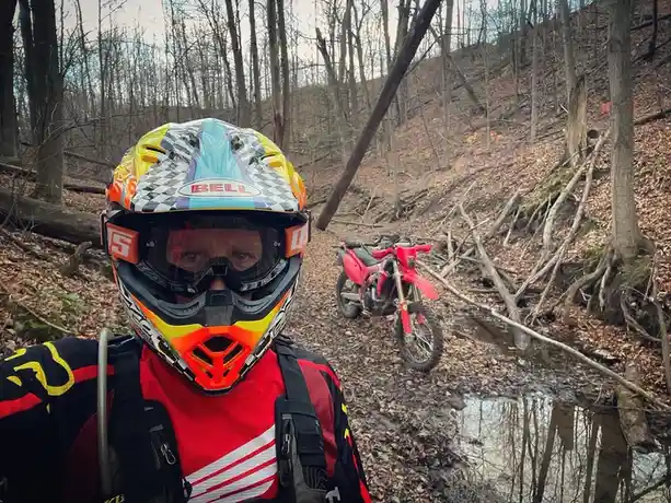 A person wearing a colorful Bell motocross helmet and goggles stands in a muddy forest, looking at the camera. Behind them…
