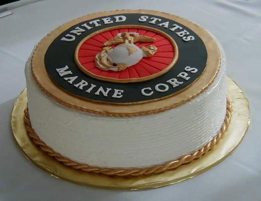 A white frosted cake with a gold rope border sits on a gold cake board. The top is decorated with the official United States…