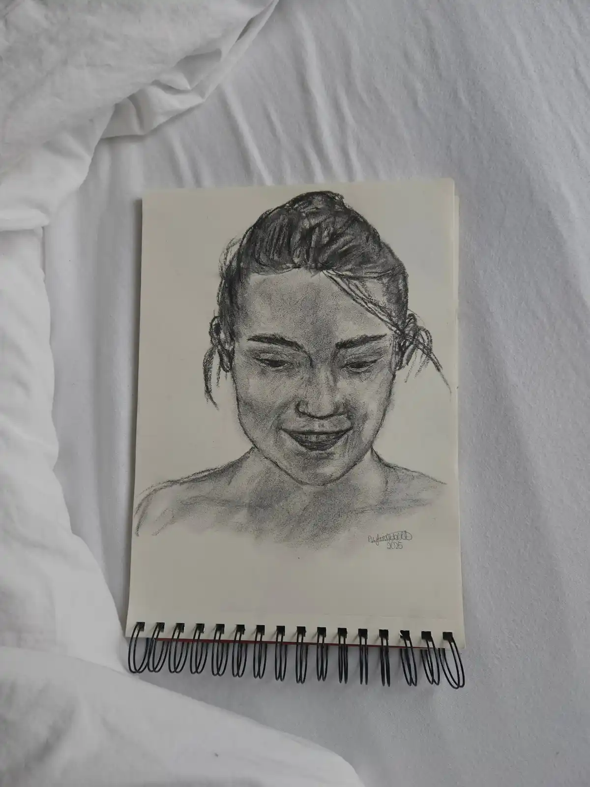 A pencil sketch of a woman's face with her hair tied up, smiling gently while looking down, drawn on a spiral-bound notebook…