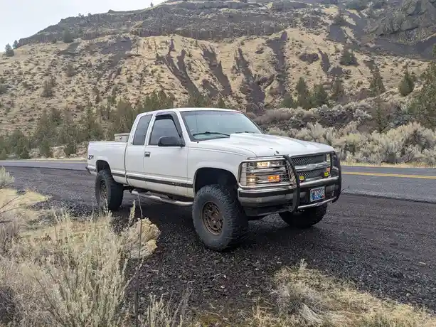 A white lifted Chevrolet pickup truck with large off-road tires is parked on a gravel shoulder next to a paved road, with a…