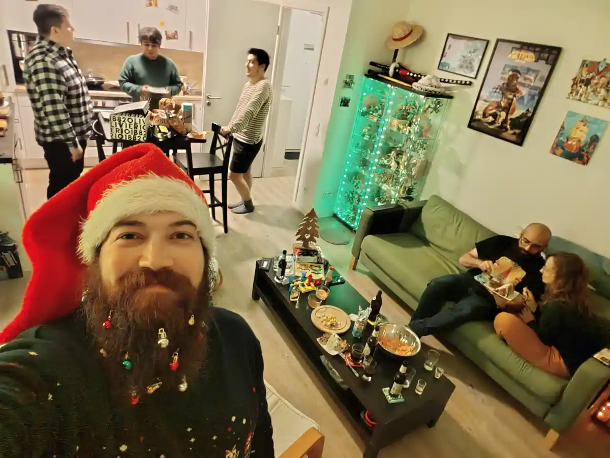 A man with a long beard decorated with Christmas ornaments and wearing a red Santa hat takes a selfie in a living room. In…