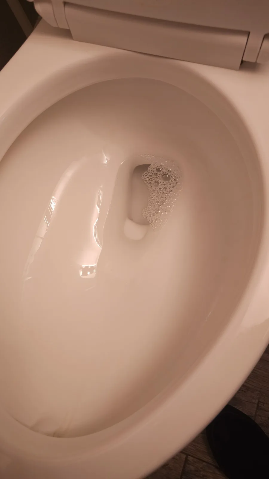 A close-up photo shows the inside of a clean, white toilet bowl with water and bubbles around the drain, the lid is up, and…