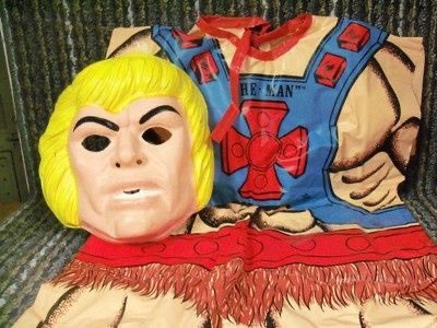 A large, worn He-Man costume mask with blonde hair and a stern expression is laid out on a couch, next to the matching blue…