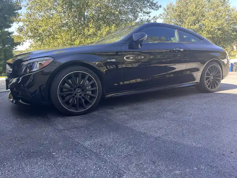 A glossy black Mercedes-Benz coupe is parked on asphalt with trees in the background. The car has a damaged front bumper and…