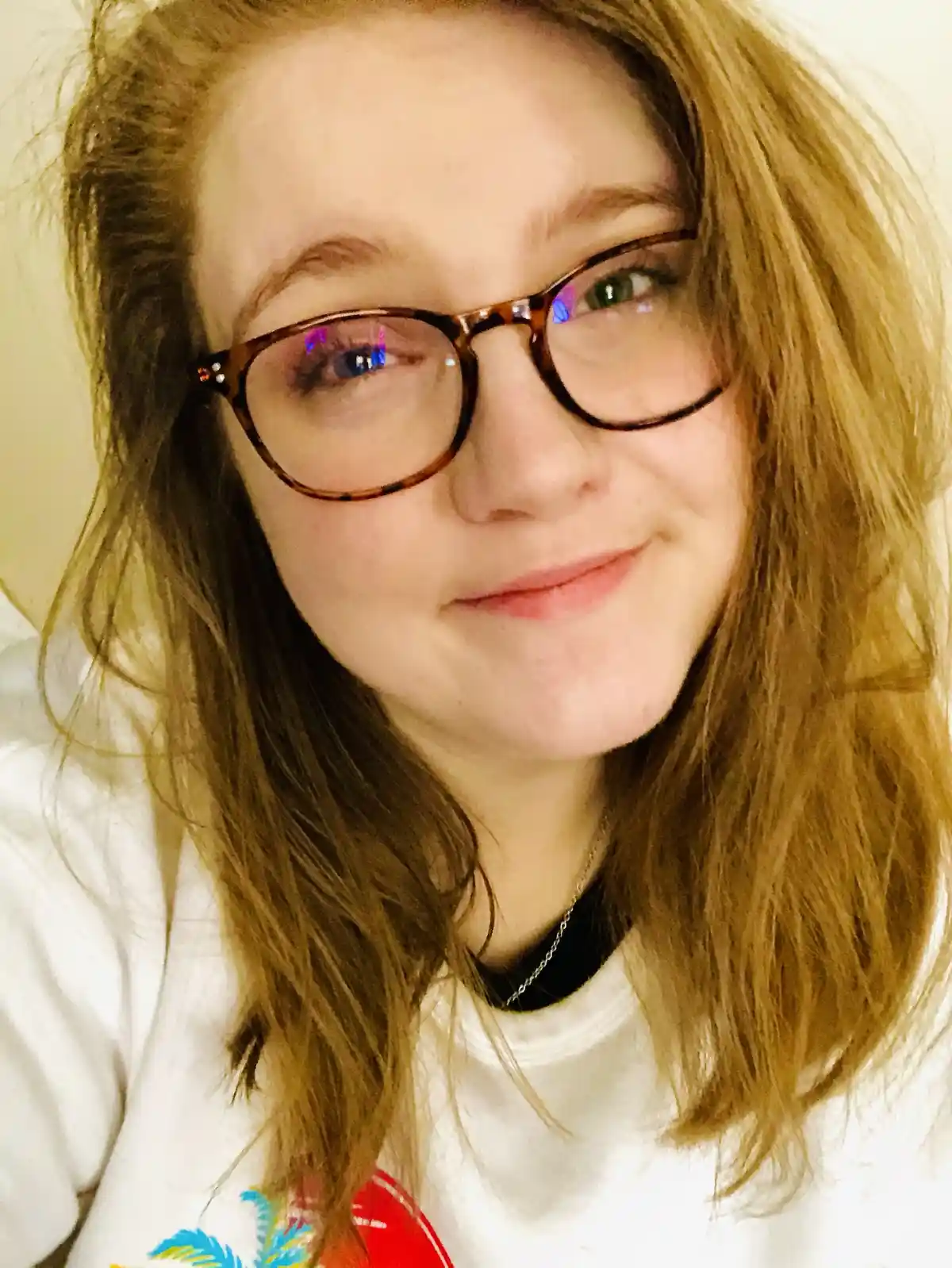 A close-up photo of a young woman with light brown hair and tortoiseshell glasses, smiling subtly while looking at the…