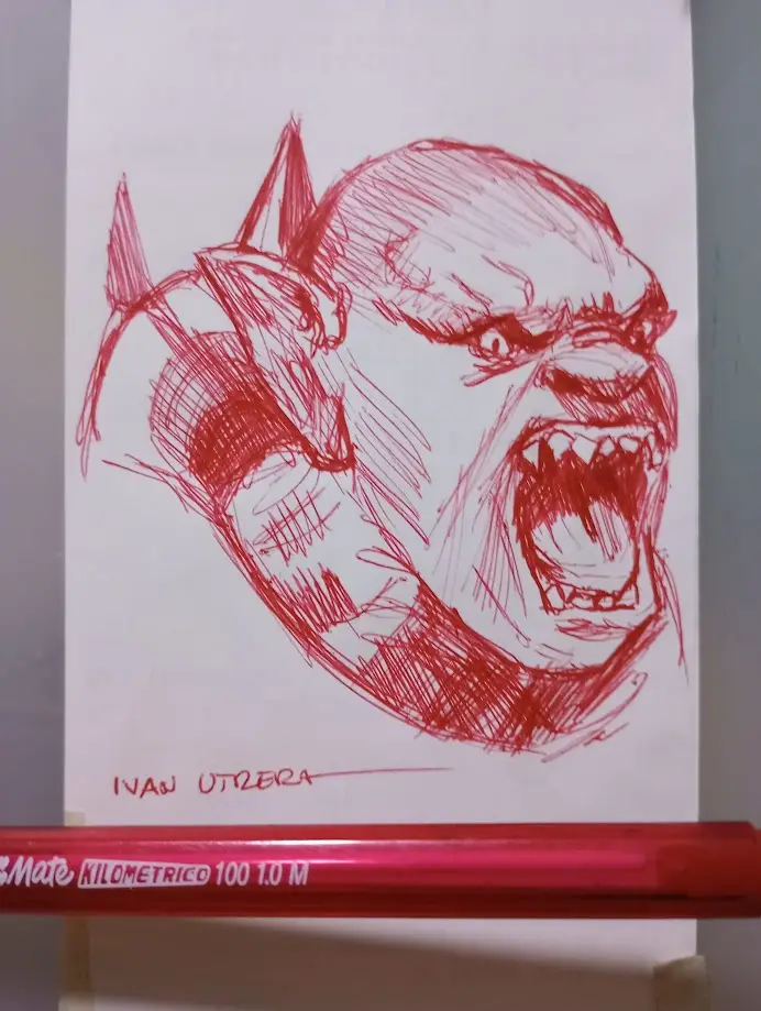 A red ink sketch on white paper depicts a fierce, snarling, horned creature with bared teeth and an angry expression. A red…
