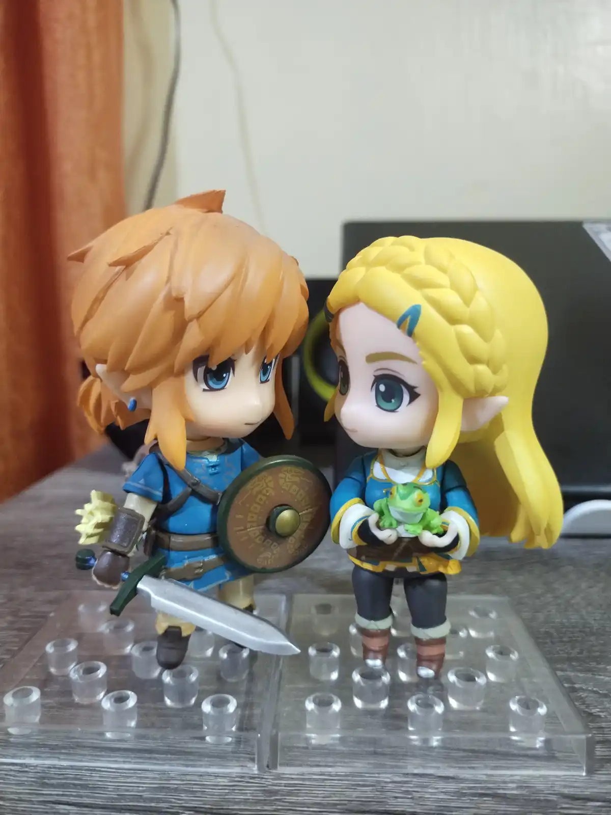 Two chibi-style figurines of Link and Zelda from The Legend of Zelda are posed facing each other on clear plastic stands…