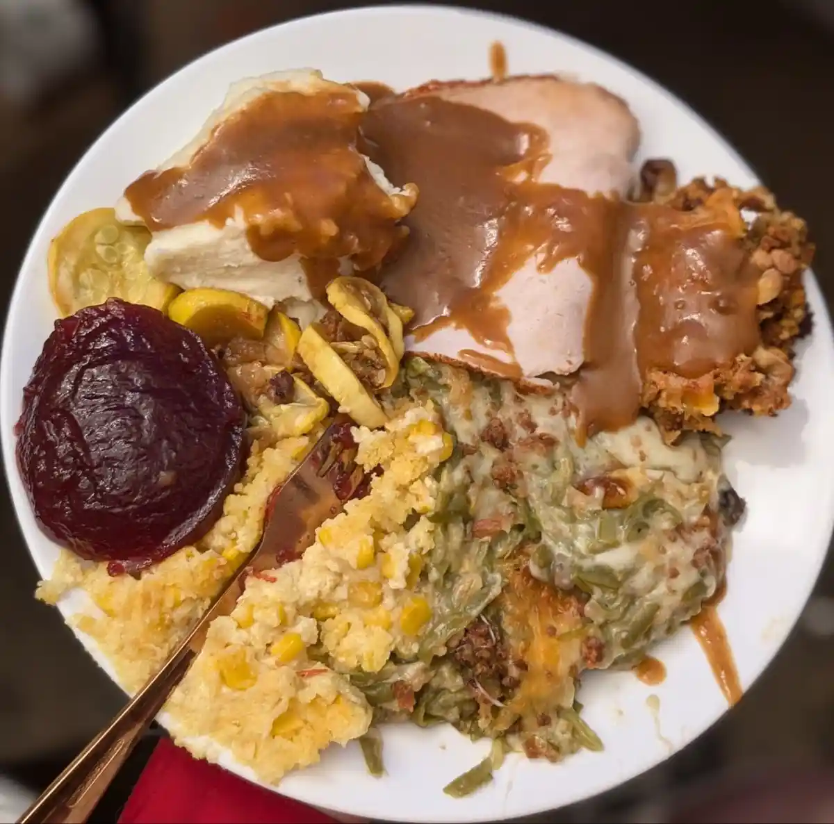 A white plate is piled high with classic thanksgiving food including turkey slices, mashed potatoes, cornbread, green bean…