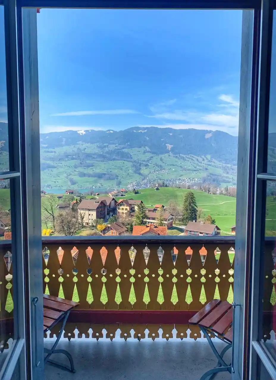 A scenic view from a balcony with ornate wooden railings and two wooden benches, looking out over a valley with green hills…