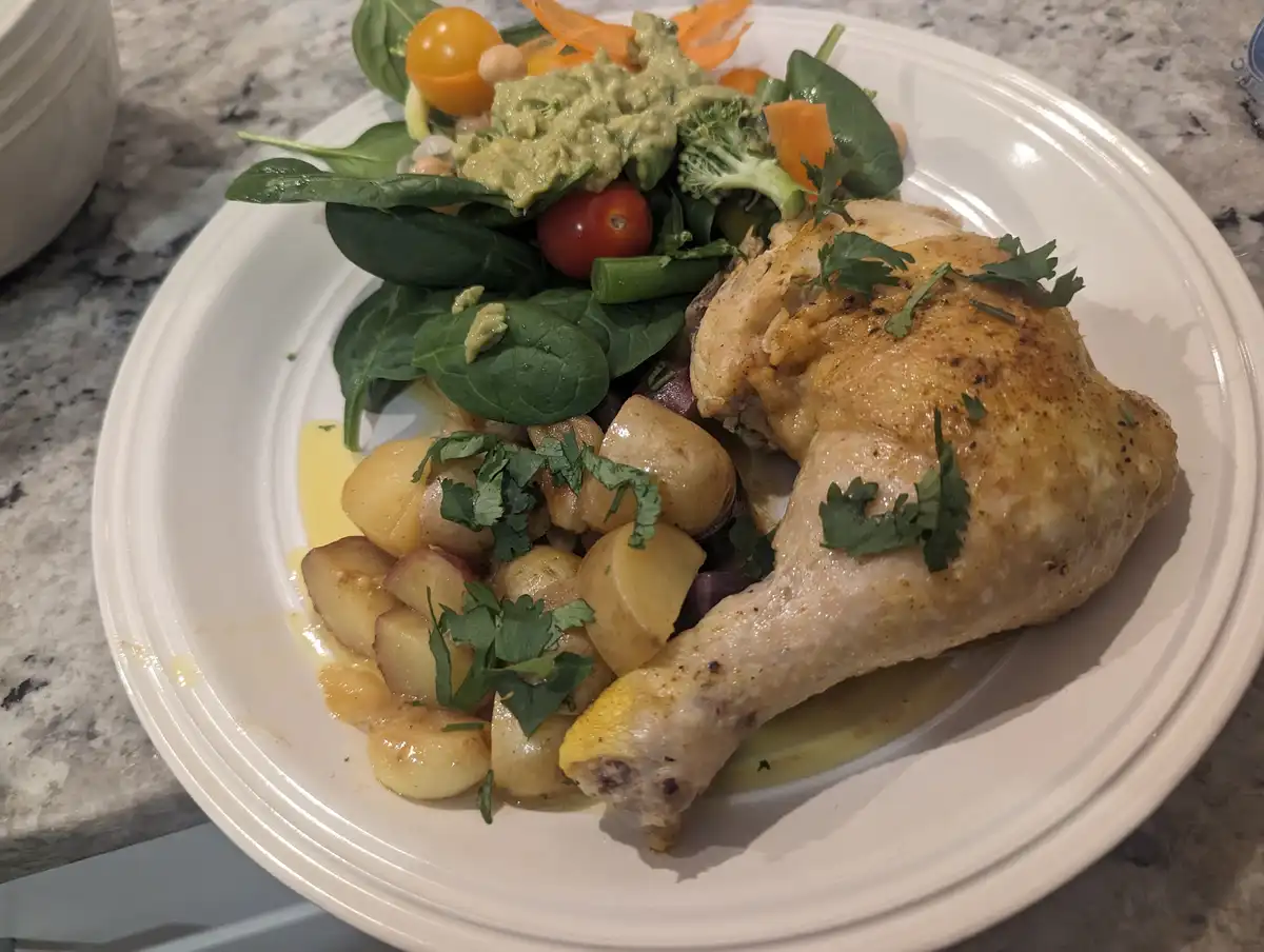 A white plate holds a roasted chicken leg, roasted potatoes, and a green salad with avocado, cherry tomatoes, and broccoli…