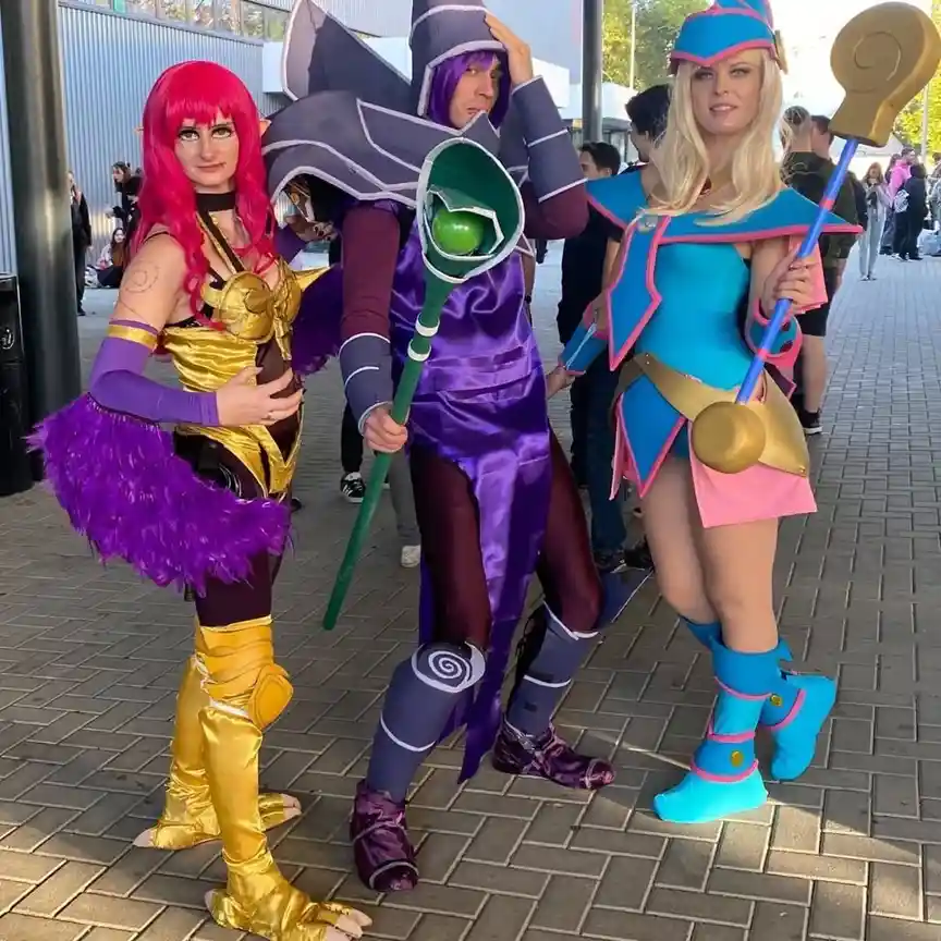 Three people in elaborate cosplay stand together outdoors on a paved walkway. The person on the left wears a gold and purple…