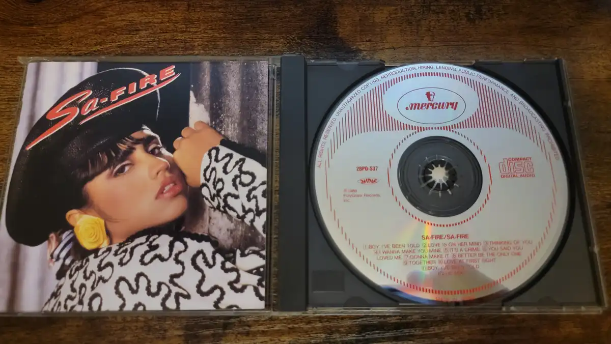 An open CD case displays the album cover of Sa-Fire, featuring a woman in a black hat and yellow rose earring, next to the…