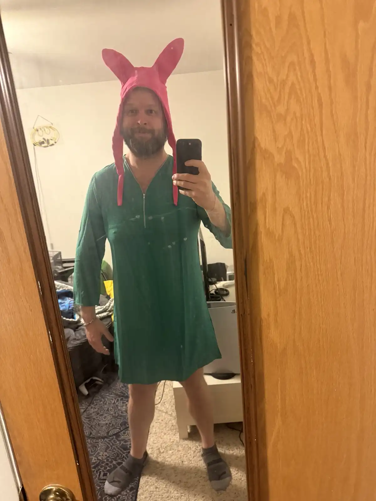 A man with a beard takes a mirror selfie while wearing a pink Beavis-style bunny hat and a green, loose-fitting dress that…