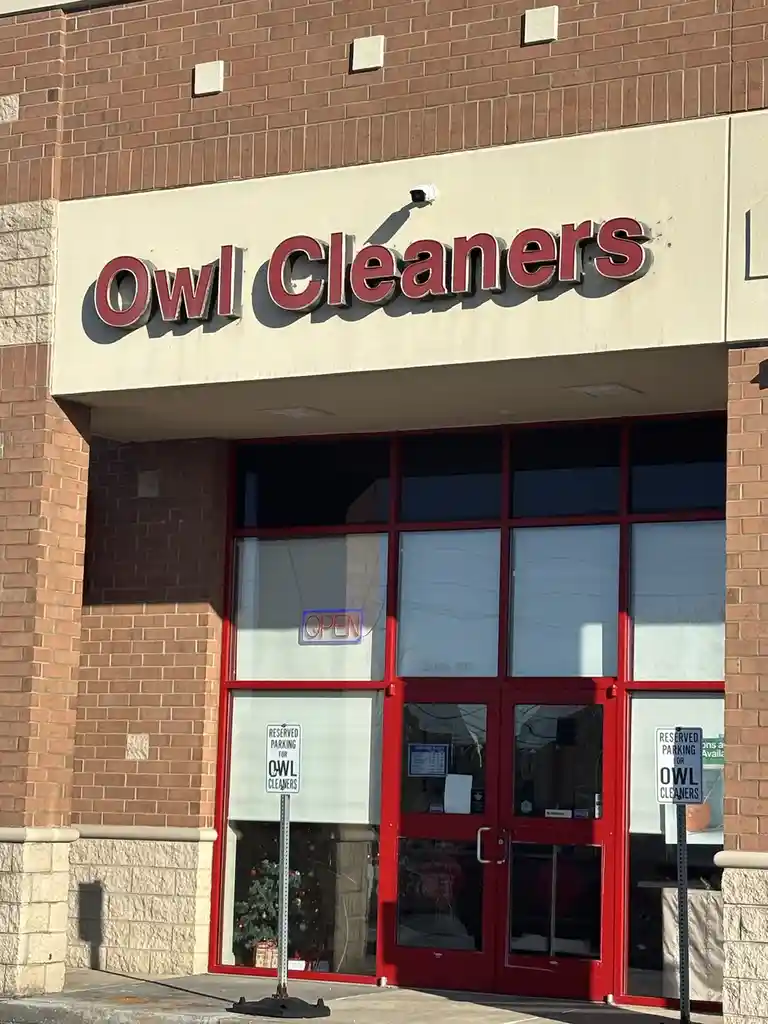 A storefront with a sign reading 'Owl Cleaners' above red-framed glass doors. An 'OPEN' sign is visible in the window, and…