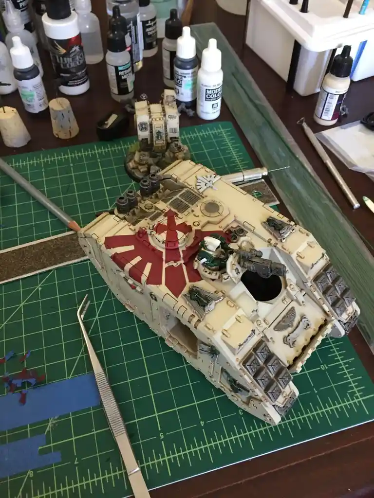 A detailed, unpainted model of a tank sits on a green cutting mat surrounded by paint bottles and hobby tools. The tank's…