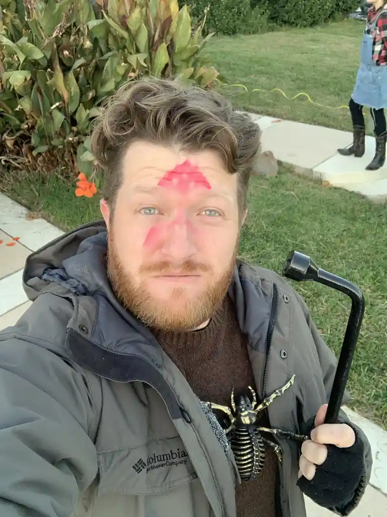 A man with a beard and blue eyes takes a selfie outdoors, his face painted with red markings resembling a spider's web…