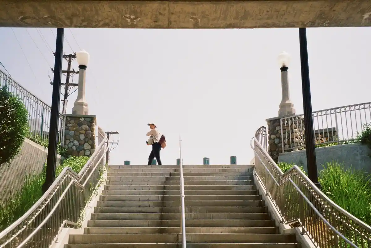 A person wearing a cap and carrying a backpack walks up a long concrete staircase framed by ornate metal railings and stone…