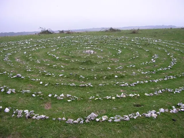 A wide-angle photo of a large, circular stone labyrinth laid out on a grassy field under an overcast sky. The labyrinth is…