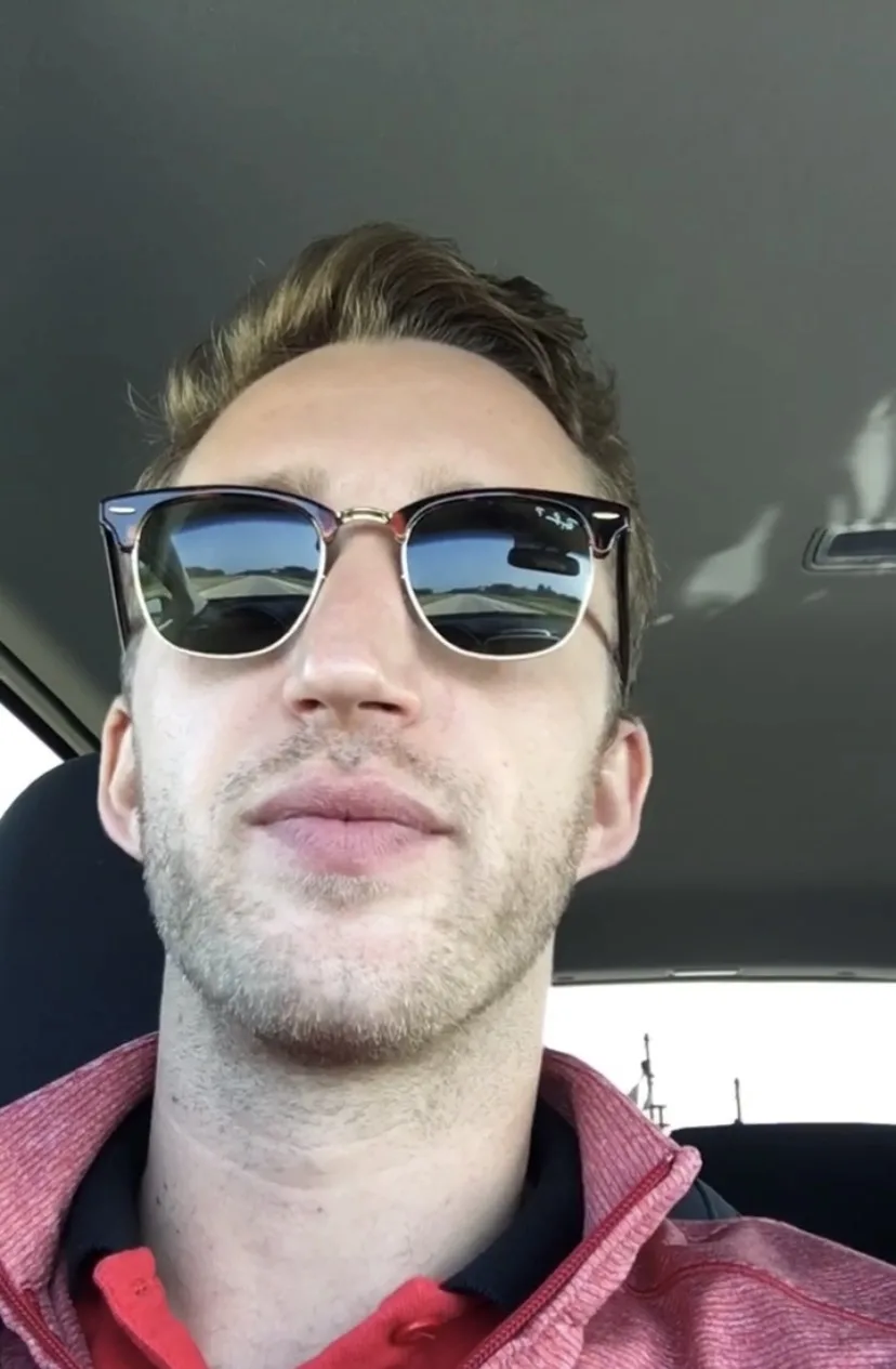 a man with light brown hair and stubble sits in a car wearing large, dark-lensed sunglasses with tortoiseshell frames. He…