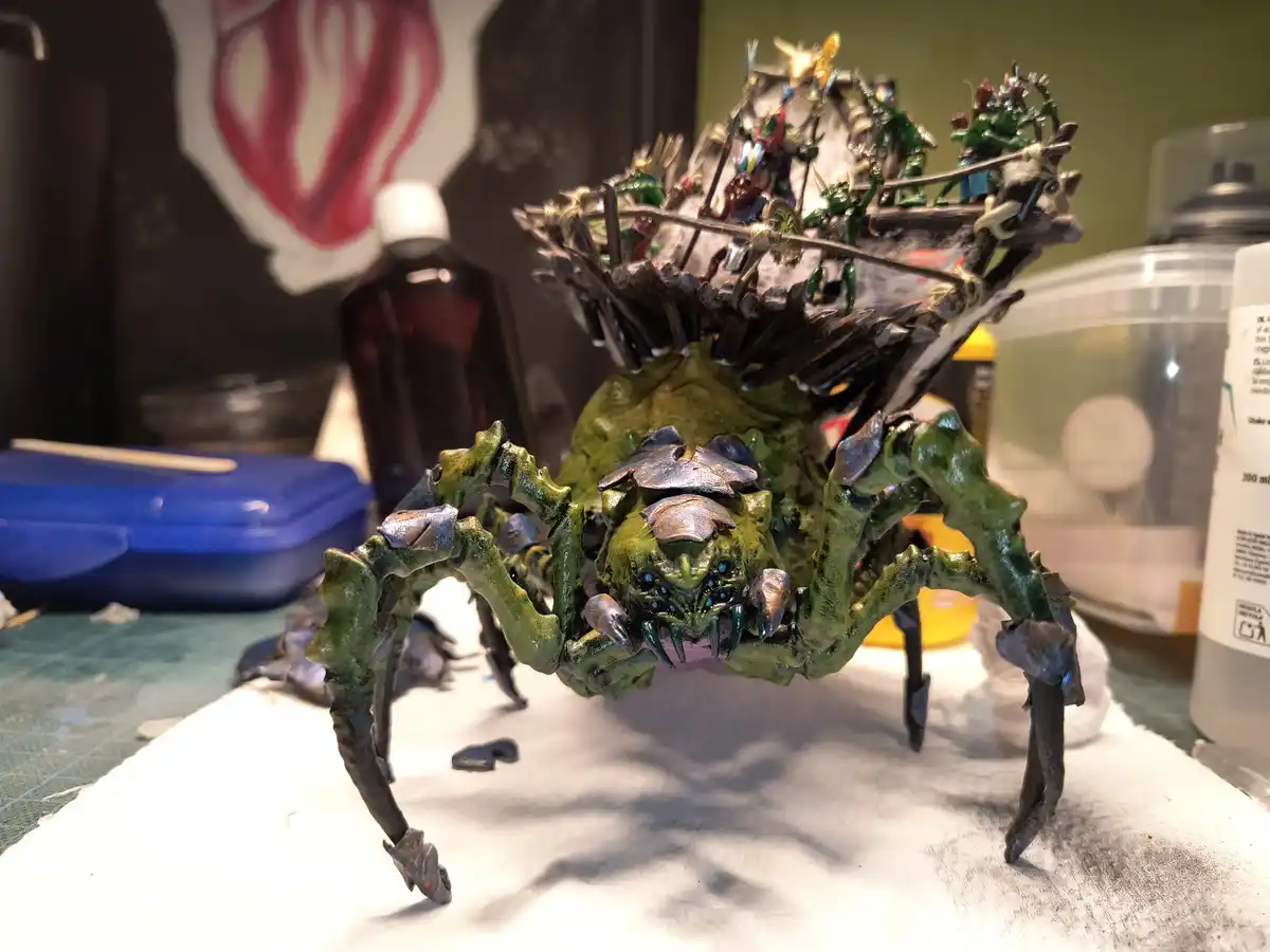 A highly detailed, green-painted miniature spider model with a menacing face and metallic claws stands on a white surface…