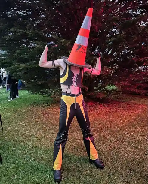 A person wearing a bright orange traffic cone on their head, flexing their biceps in a muscle pose. They are dressed in a…