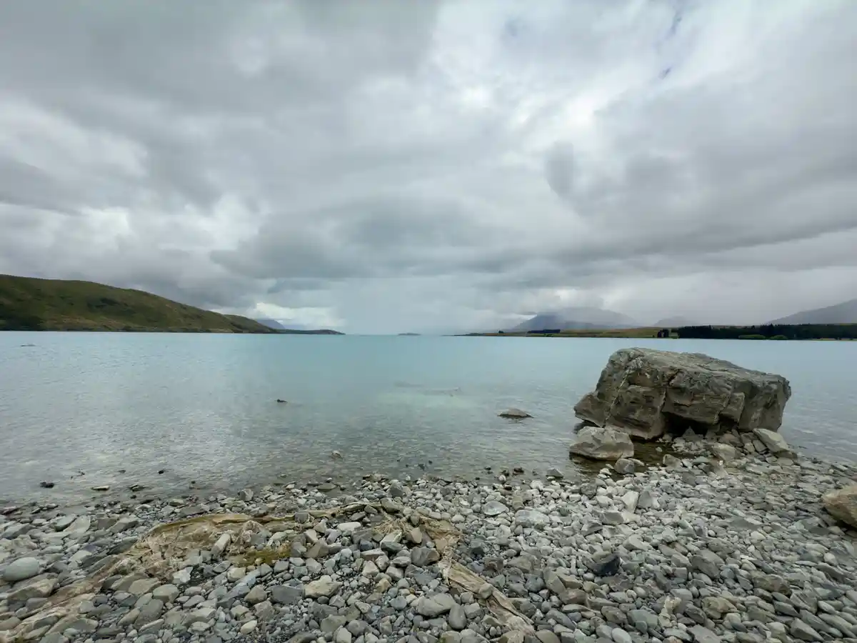 A serene, overcast landscape featuring a calm lake bordered by rolling hills and a rocky shoreline with a large boulder. The…