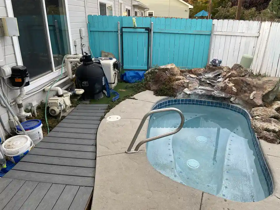 An outdoor hot tub with blue tiled edges and a metal ladder is surrounded by a concrete deck and a wooden boardwalk. Pool…