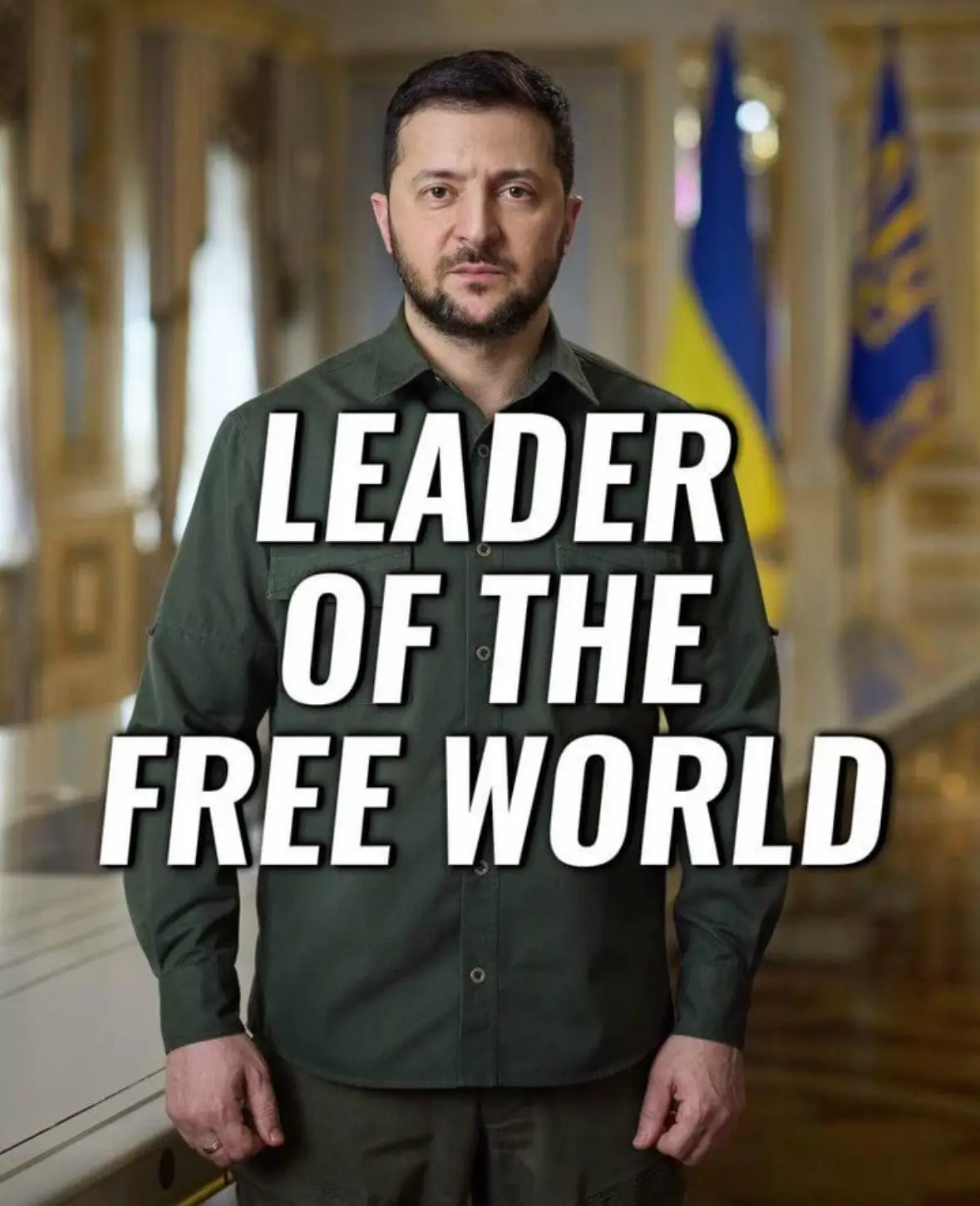 A man with a beard and short dark hair, wearing a dark green button-up shirt and matching pants, stands in a formal room…