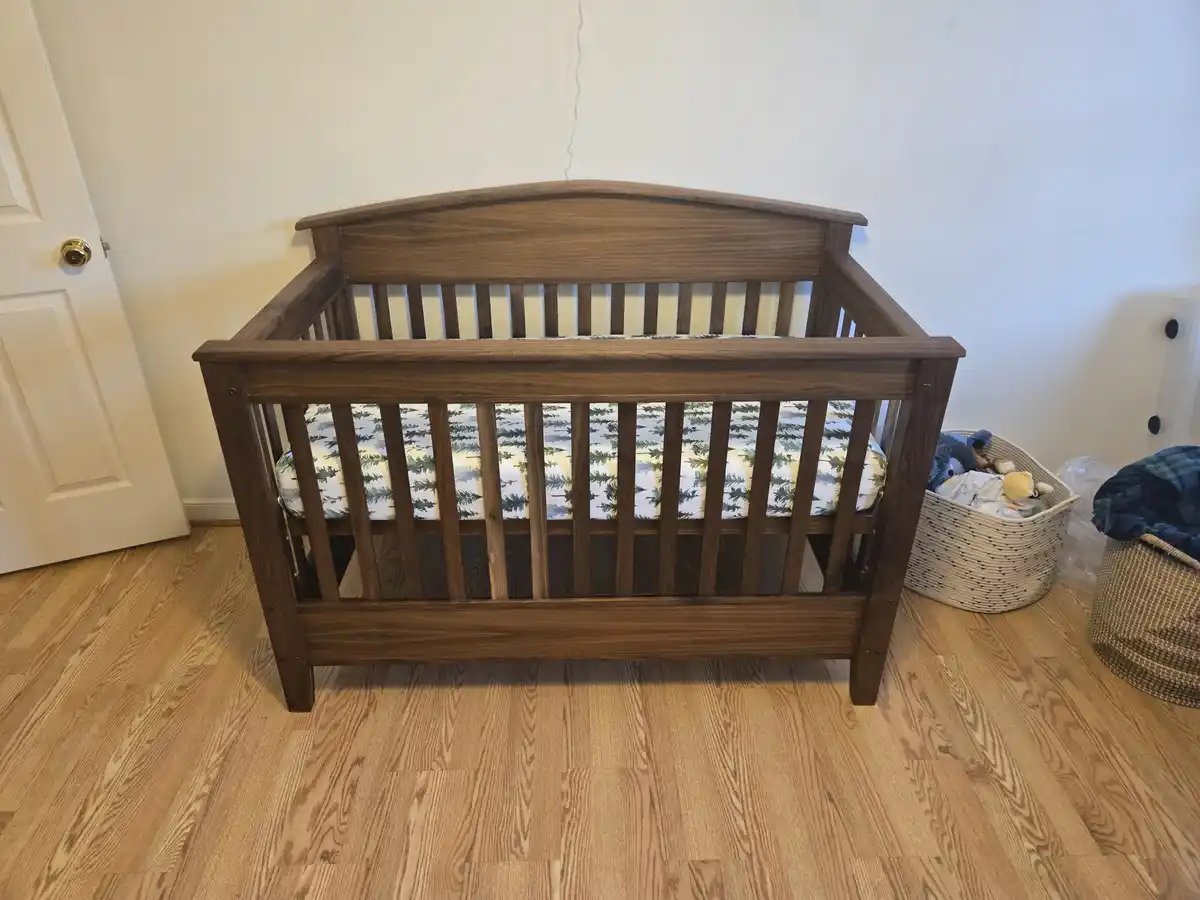 A dark wood baby crib with a forest-patterned mattress is centered on a light hardwood floor in a room with white walls and…