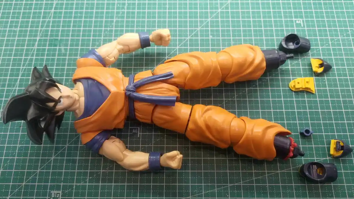 A broken Dragon Ball Z action figure of Goku lies on a green cutting mat, with his arms and legs detached and scattered…
