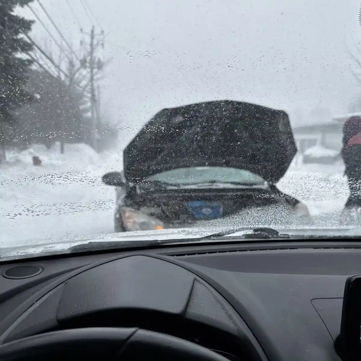 A view from inside a car during a heavy snowstorm, looking out at another car with its hood open. The windshield is covered…
