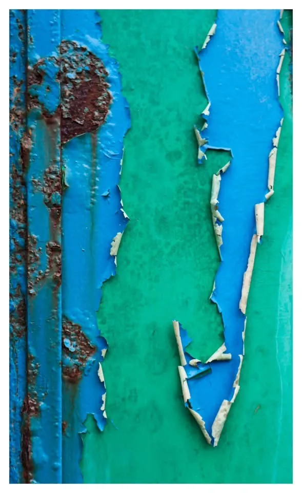 A close-up photo of weathered metal with peeling blue paint revealing a green layer underneath and rust stains on the left…