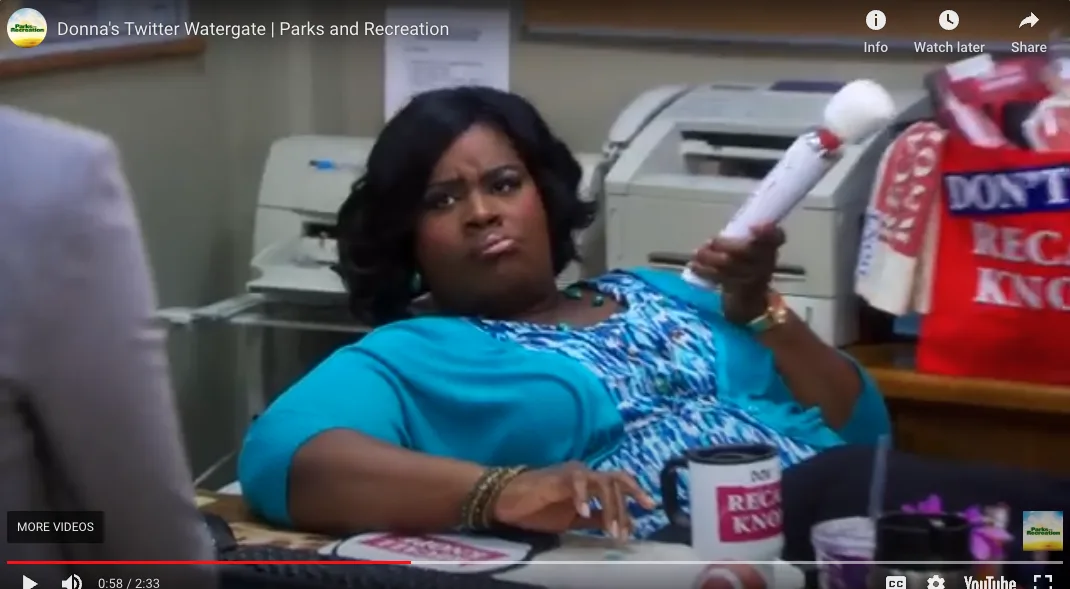 A woman in a blue top sits at a desk holding a white foam finger, looking annoyed. A mug and a red bag with the text 'DON'T…