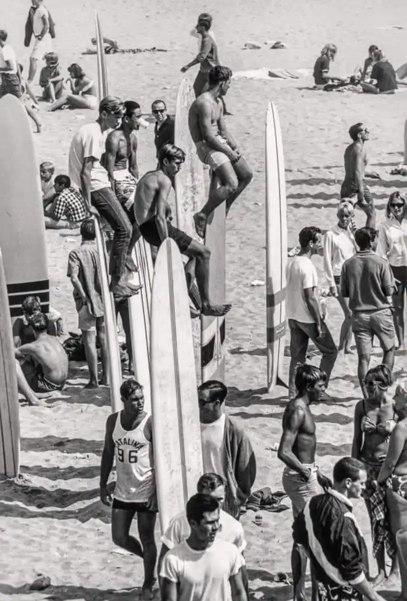 A black-and-white photo shows a crowded beach scene with numerous surfers and beachgoers. Several men are perched atop tall…