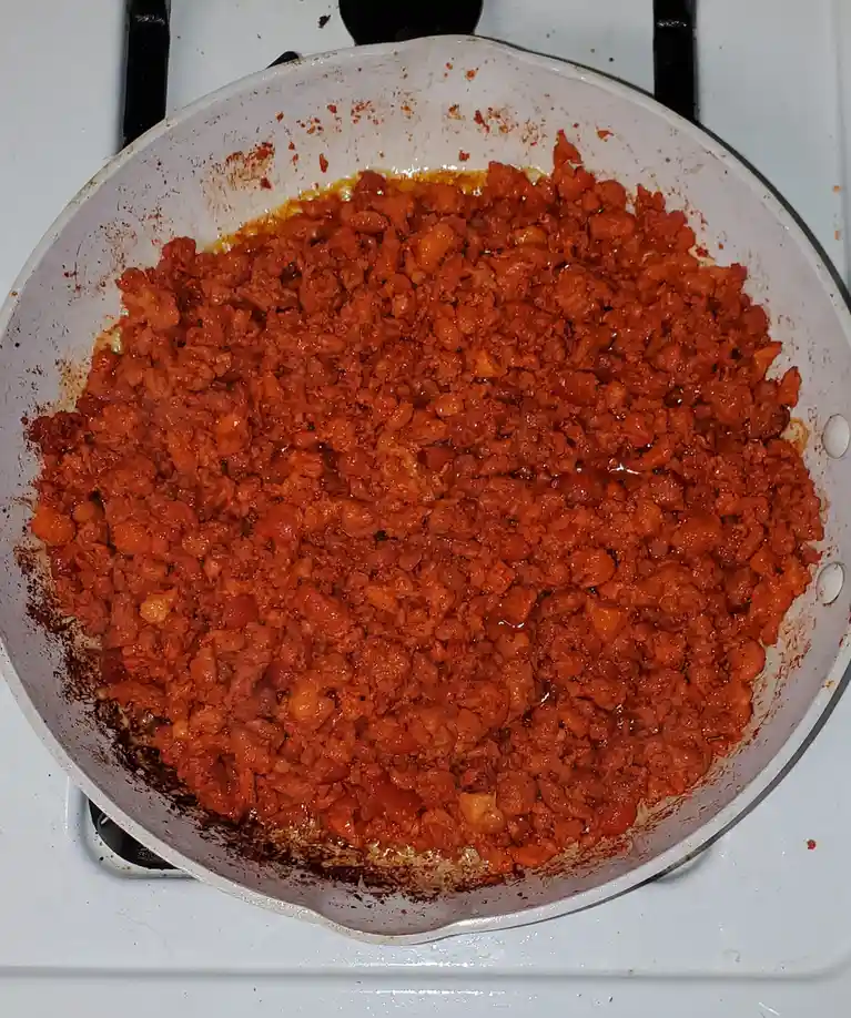 A close-up photo shows a white frying pan on a stovetop filled with a reddish-brown, chunky mixture that looks like cooked…