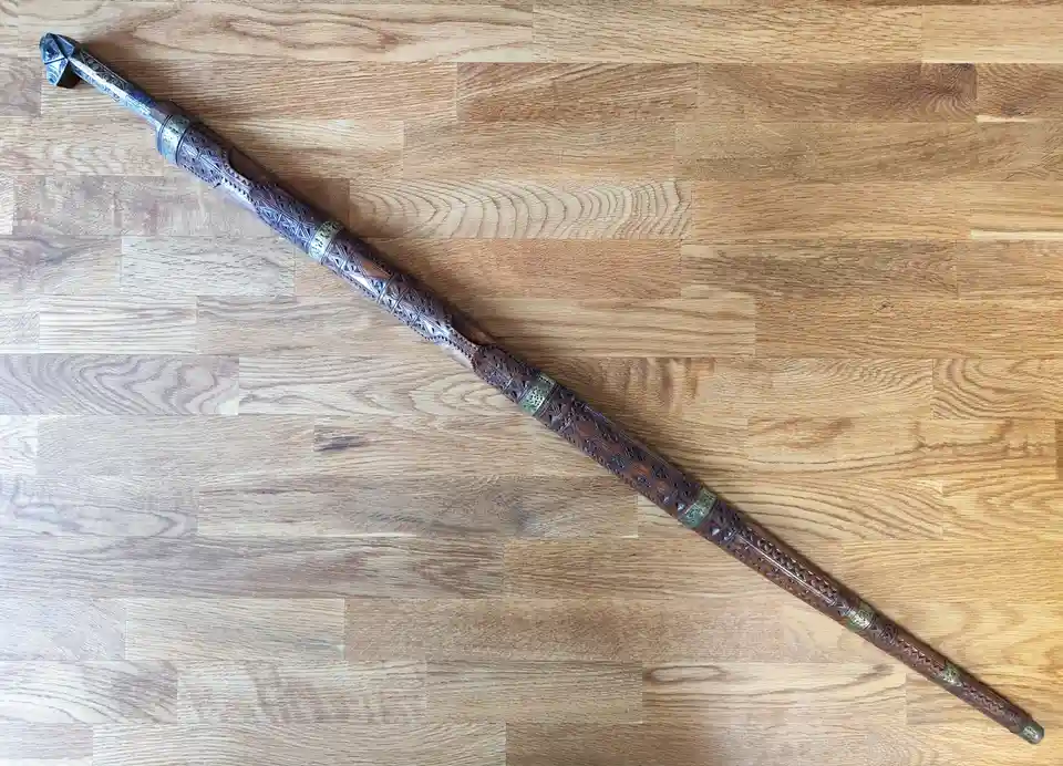 a long, intricately carved wooden walking stick with metal accents lies diagonally across a light brown wooden floor. The…