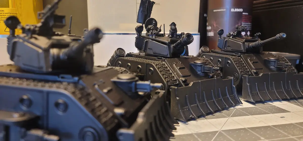 A photo of three detailed black miniature tanks with bulldozer blades attached to their fronts, displayed on a checkered…