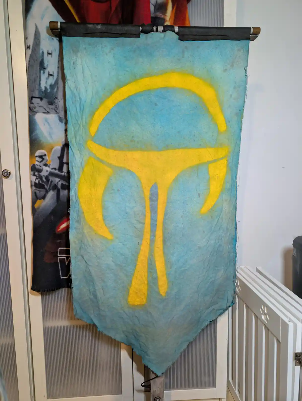 A handmade, light blue fabric banner with a yellow Mandalorian symbol painted on it hangs from a rod in a room. To the left…