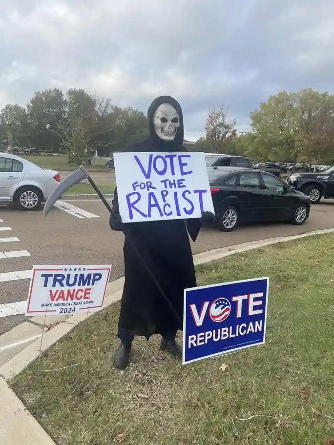 A person dressed as the grim reaper, holding a scythe and a handmade sign that reads 'VOTE FOR THE RACIST', stands in a…