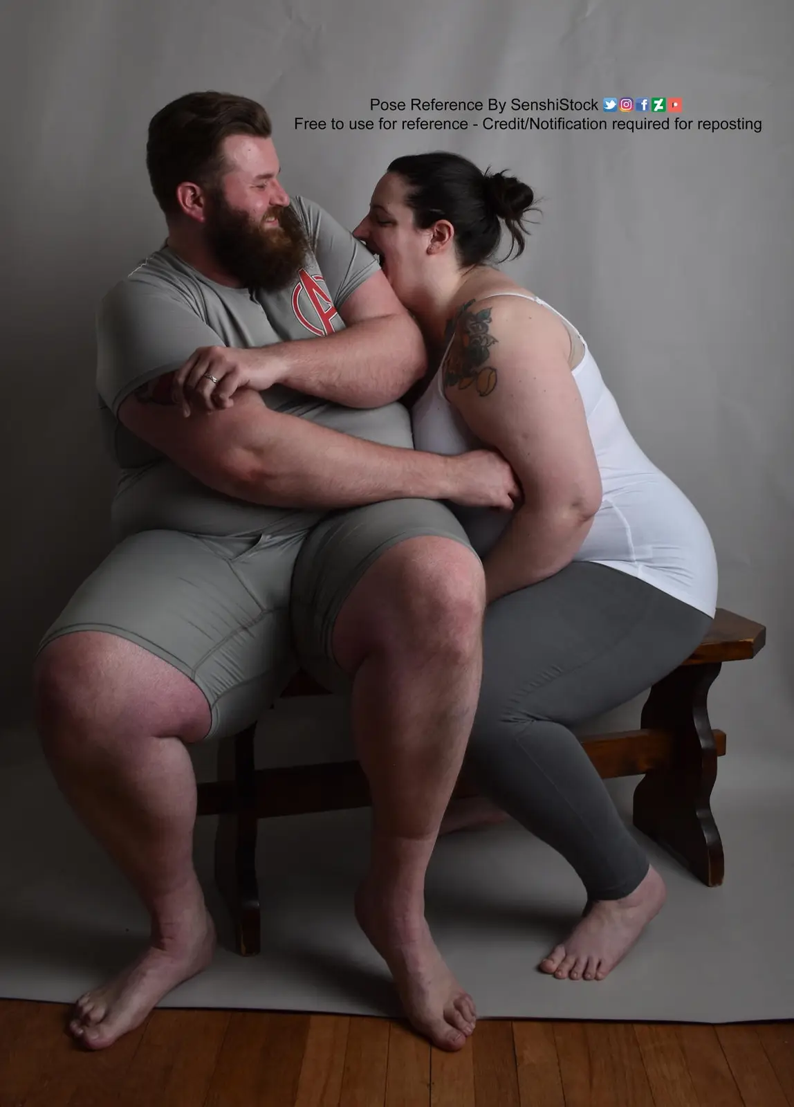 A man and woman are sitting closely together on a wooden bench against a plain gray backdrop, both smiling warmly at each…