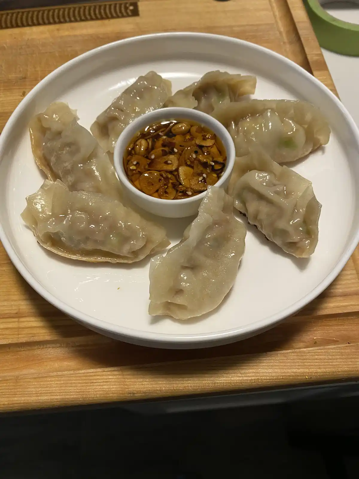 A white plate holds seven translucent, pan-fried dumplings arranged around a small bowl of dipping sauce containing peanuts…