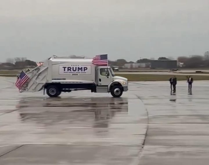 A white garbage truck with 'TRUMP 2024' and 'MAKE AMERICA GREAT AGAIN' written on its side is parked on a wet tarmac…