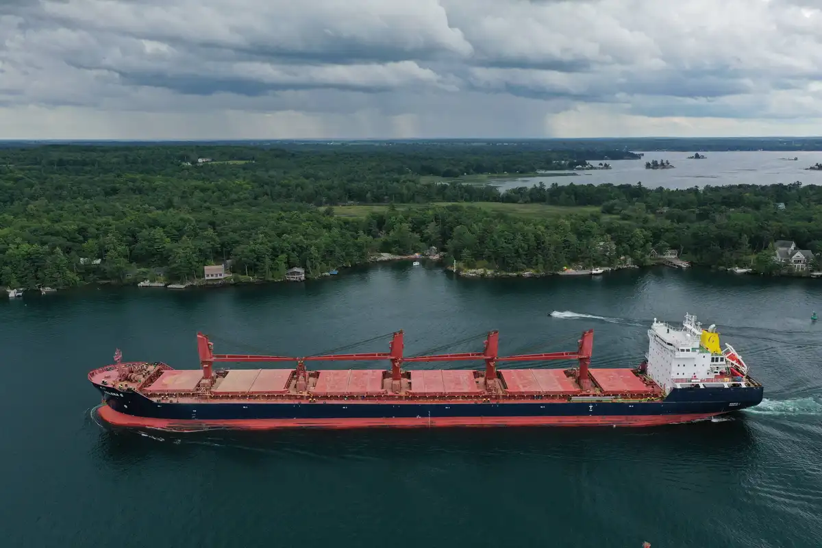 An aerial photo shows a large red and blue cargo ship sailing through a narrow waterway surrounded by dense green forests…