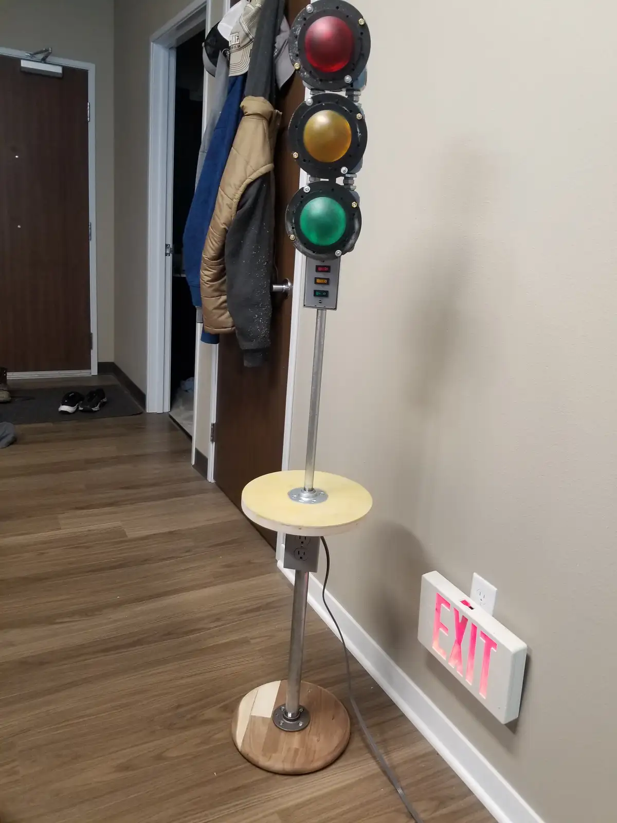 A homemade traffic light with red, yellow, and green bulbs mounted on a wooden stand in a hallway, with the red light…