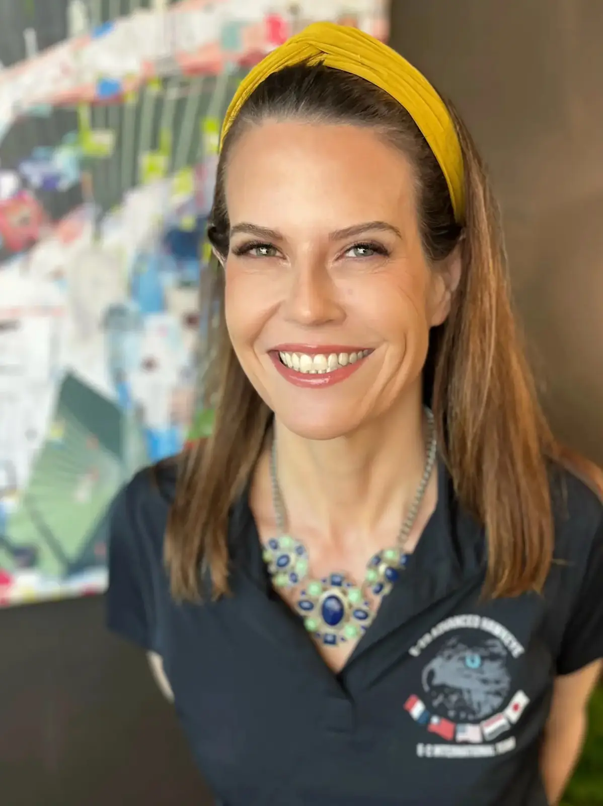 a woman with brown hair and a yellow headband smiles while wearing a dark polo shirt with a logo and a statement necklace…